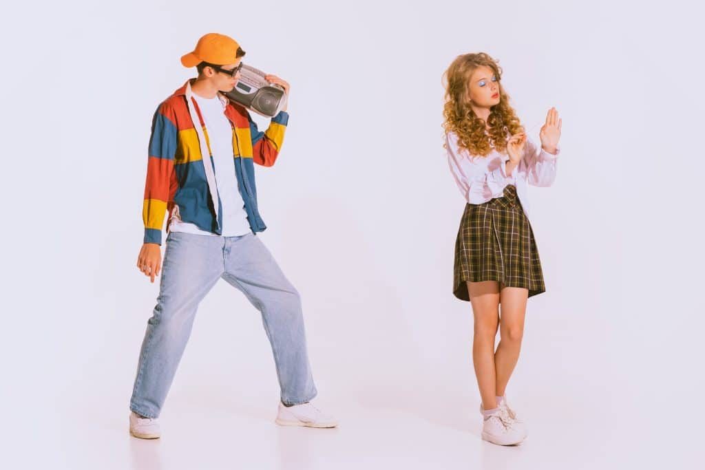 Young man dressed in colorful windbreaker and holding a CD player looking at young girl dressed in casual preppy aesthetic - corporate fashion