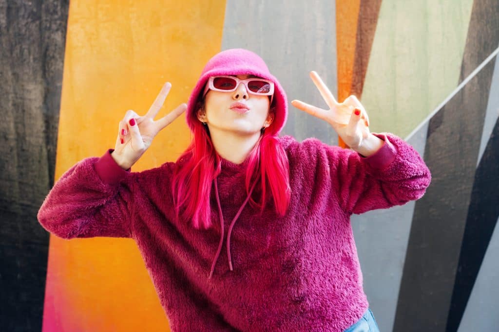 Woman with opnk hair, hat, sunglasses, and sweater posing in front of an orange and grey background.