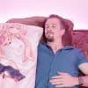 A man lies on a bed. In his arms is a body pillow with the image of a young girl wearing a school uniform with visible panties.