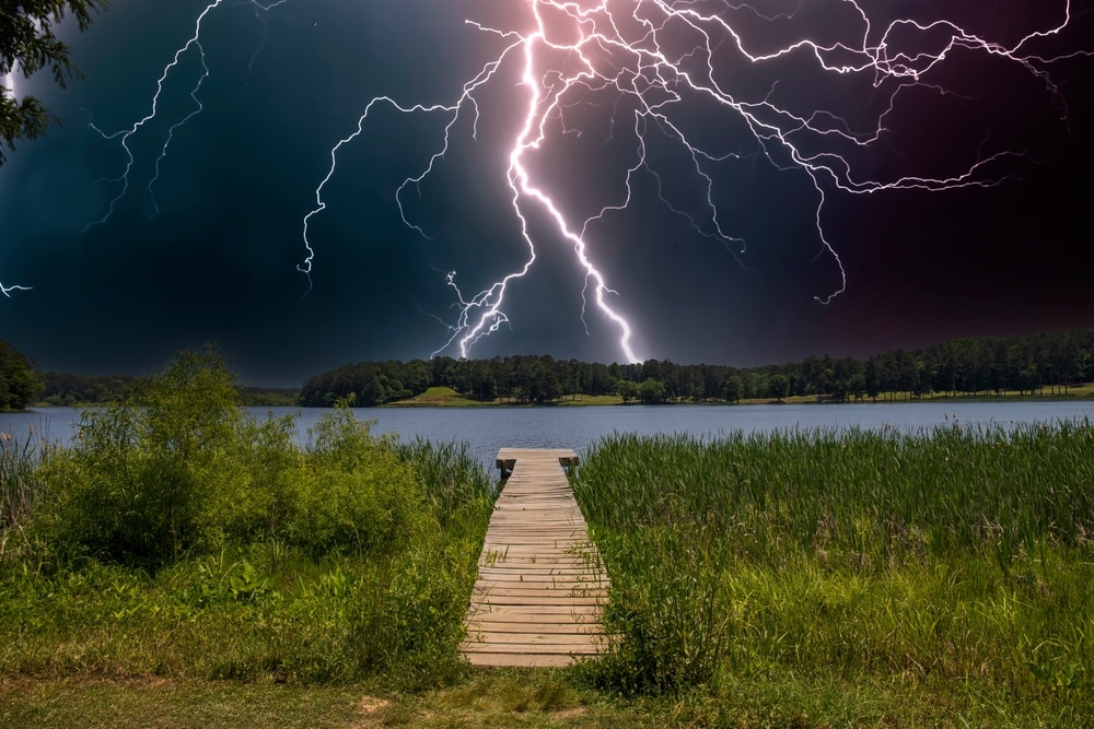 Lightning strikes in a stormy sky. Scenery includes trees, a lake, and a wooden dock.