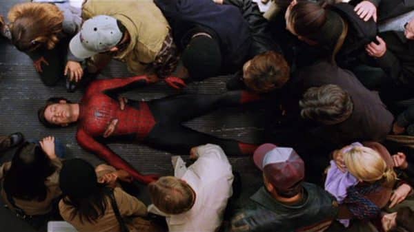 An unmasked Spider-Man lies on a train floor as people surround him