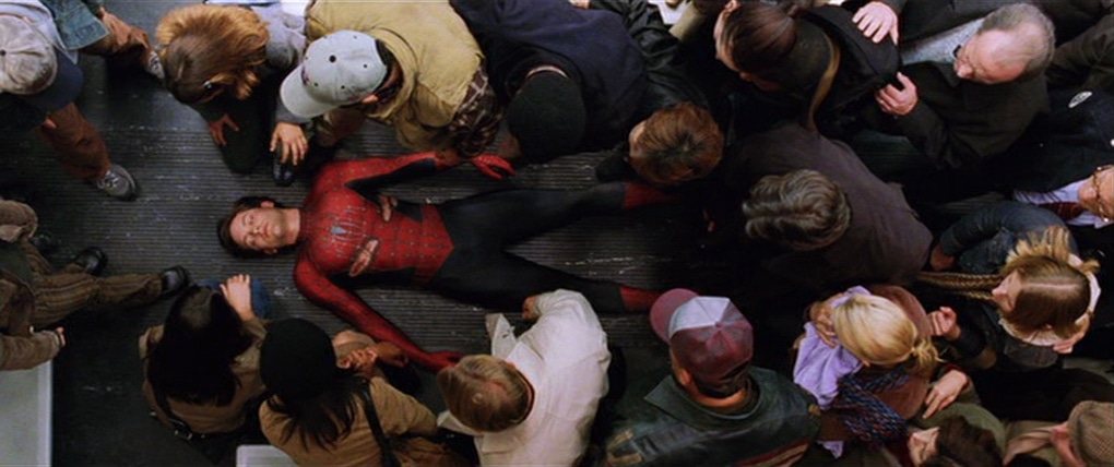 An unmasked Spider-Man lies on a train floor as people surround him