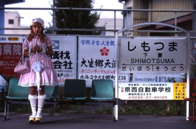 Momoko stands in front of a Japanese bus station. She is wearing a pastel pink Lolita outfit with clean white socks and a parasol in gloved hands.