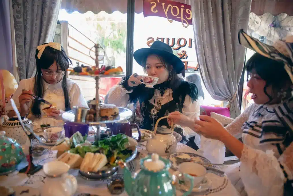 Multiple women in Lolita fashion are sat around a table drinking tea. In front of them are fancy teacakes.