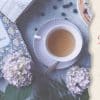 photo of table from Birds Eye view. The table has a lilac cloth on the surface and has small dark berries scattered on the top. Also on the table is a purple cup and saucer filled with tea, purple heads of hydrangea flowers, a copy of Pride and Prejudice, and some green leaves. Next to the table is a vertical beige banner with the articles title written in brown and pink font