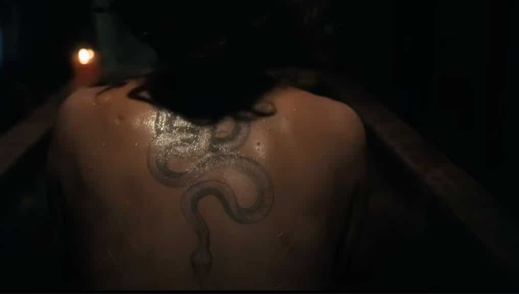 IN a dark room, a lady sits with her wet back to us, revealing her snake tattoo.