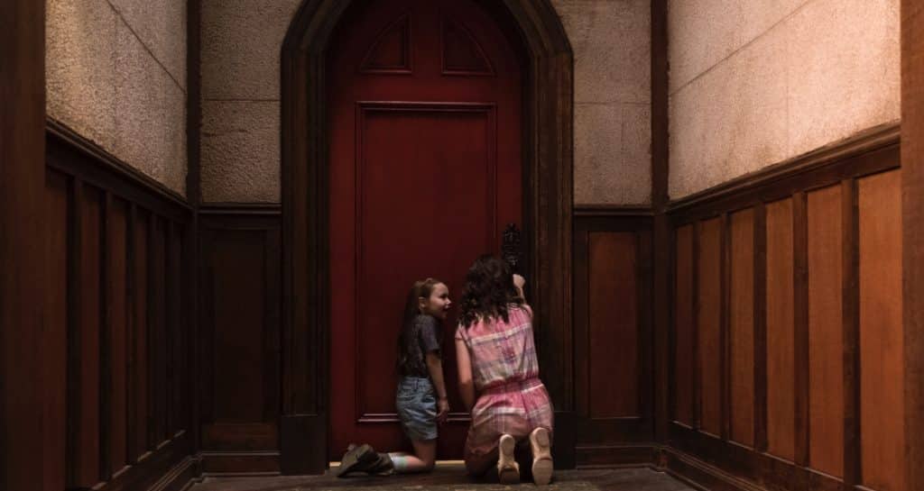 Two young girls crouch at a red door, attempting to open it.