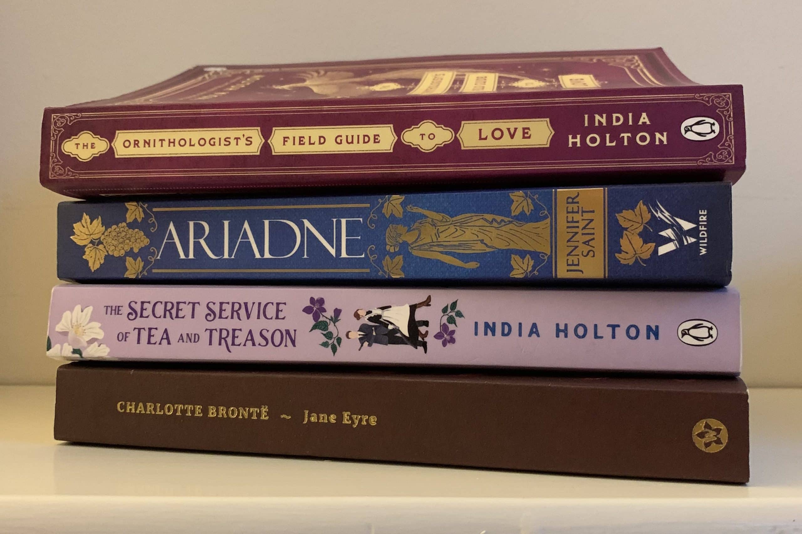 Photo of four books stacked one on top of each other with spines facing out. Book on top is a violet spine with golden banners. in the banners is the title and author's name 'the ornithologists field guide to love by India Holton'. Book below is blue with gold embossed leaves and a greek statue of a woman 'Ariadne' by Jennifer Saint. Book below is lilac 'Tje secret service of Tea and Treason' by India Holton. Book on bottom is a maroon book with the title and author's name in gold 'Jane Eyre' by Charlotte Bronte