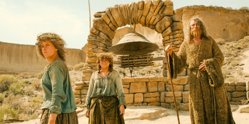Still from Rings of Power: Nori, Poppy and 'The Stranger' standing in a desert in front of a well