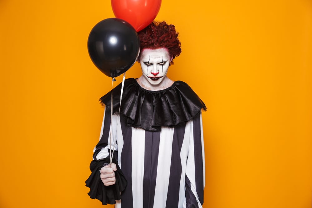 Man dressed as a creepy clown, wearing black and white stripes and holding balloons. Yellow background.