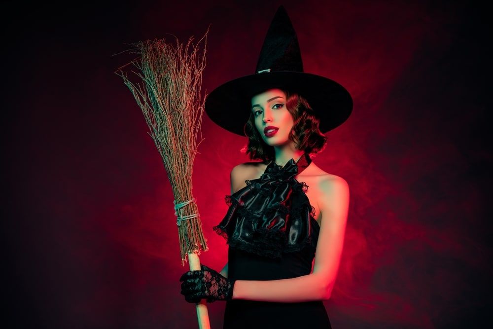 A woman in a timeless witch costume, holding a broom. Isolated, dark red background.