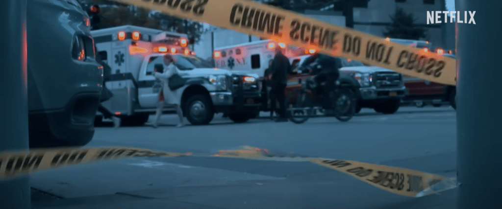 Crime scene tape flying in the wind in front of ambulances and pedestrians.