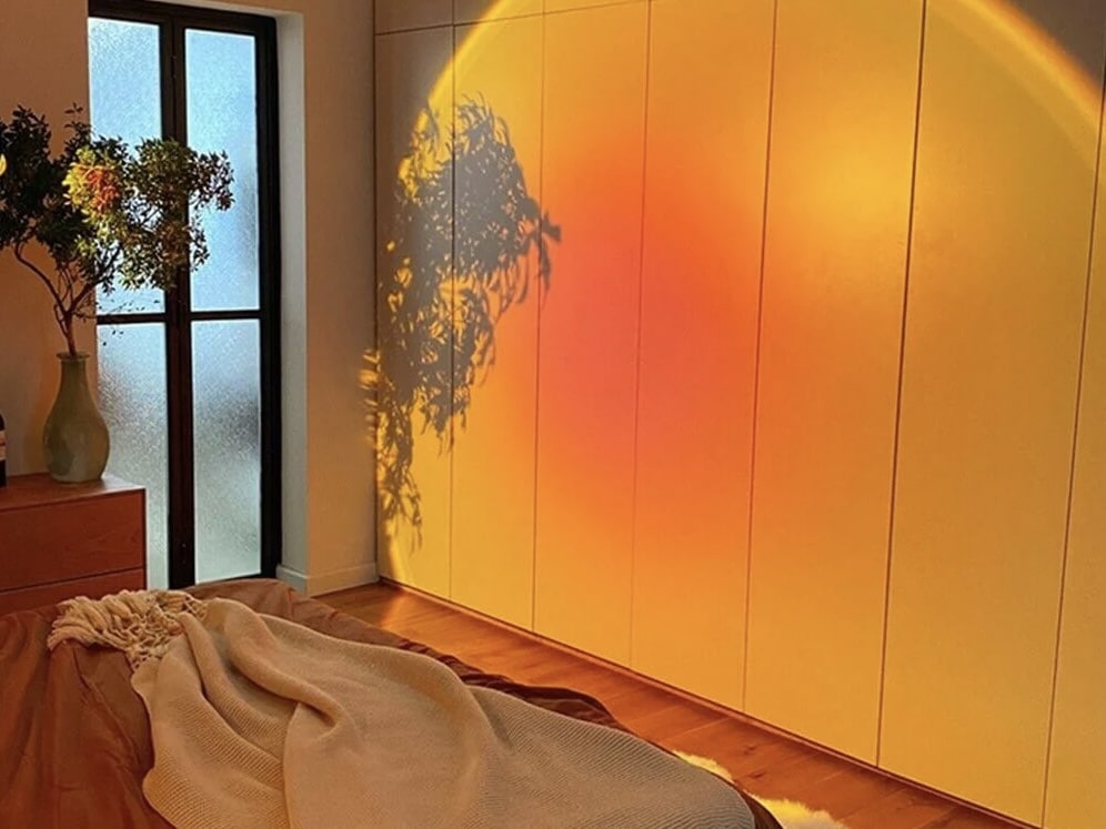 Cozy bedroom with warm mood lighting projecting a glowing sunset-like effect on the wall. A bed with soft blankets and throws occupies the foreground, while a vase with greenery sits on a dresser near a frosted glass window. The lighting casts subtle shadows of the plant, enhancing the serene and inviting atmosphere. Good example for Vitamin D use to help combat seasonal depression