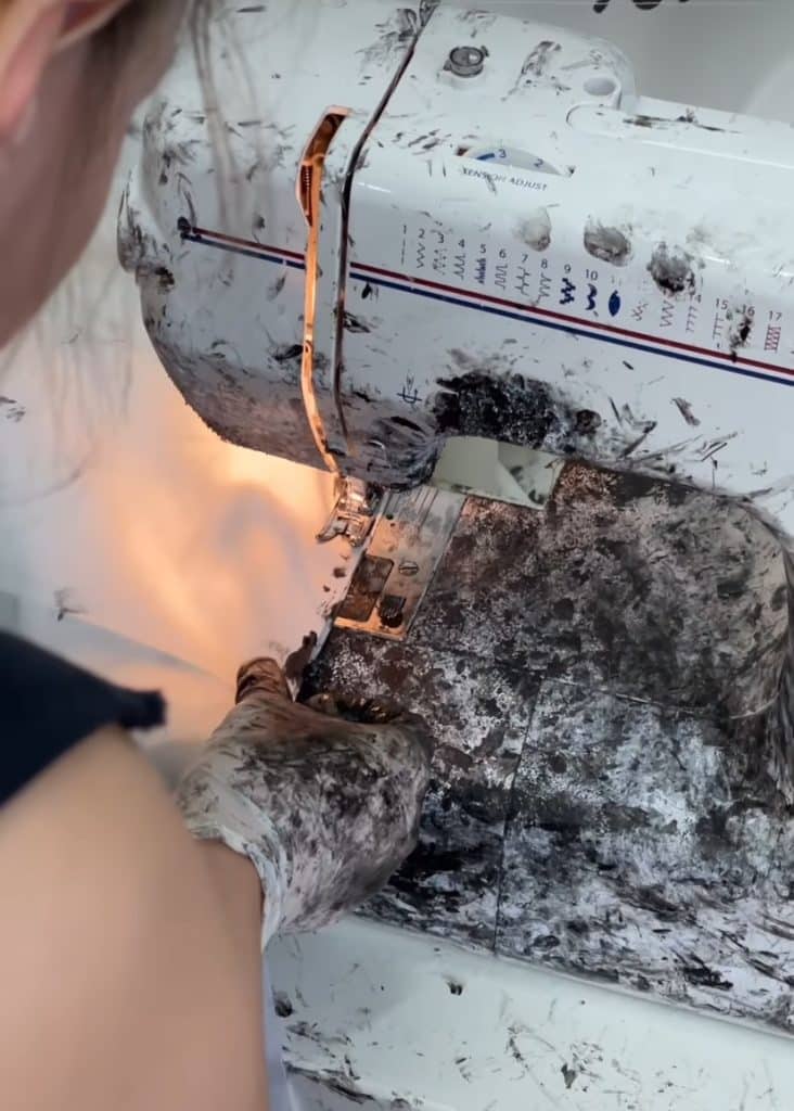 A photo of a worker sewing a white garment with black paint on their hands. Each touch is left visible on the garment thanks to the paint.