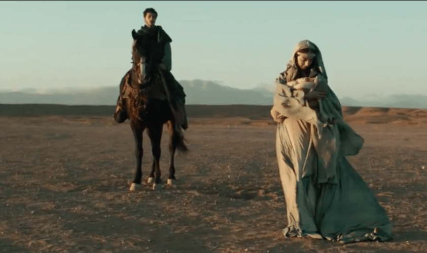 a man stands on the horse while mary walks and carries a baby. the desert is all around them.