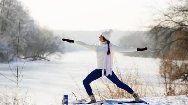 Photo of a woman doing yoga in the snow with a hat and scarf on as winter workout.