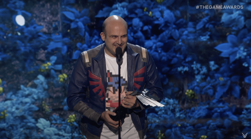 A man wearing a Zelda-inspired jacket smiles and speaks into a microphone while holding a silver trophy at the Game Awards 2024.