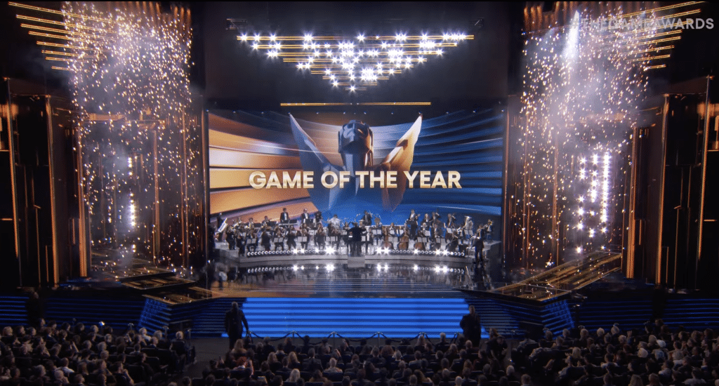 A large orchestra performs on a stage, the backdrop an image of the game awards statuette and the words "Game of the Year." Showers of fireworks fall on both sides of the orchestra.