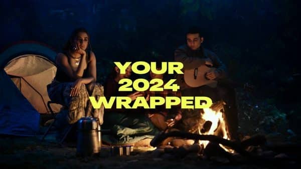 A group of friends sits around a campfire at night, enjoying a peaceful outdoor setting with a tent in the background. One person plays a guitar while others reflect and connect, symbolizing a moment of introspection. The bold text “YOUR 2024 WRAPPED” overlays the image, highlighting the theme of pre-New Year resolutions and personal growth.