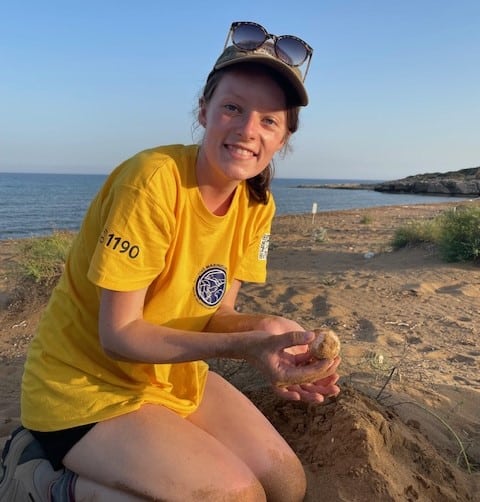 A young woman wearing a yellow volunteers top, holding a small turtle egg whilst kneeling in the sand.