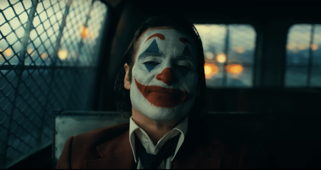 A screenshot of the trailer for "Joker: Folie À Deux," showing the Joker riding in the back of a police car in clown makeup.