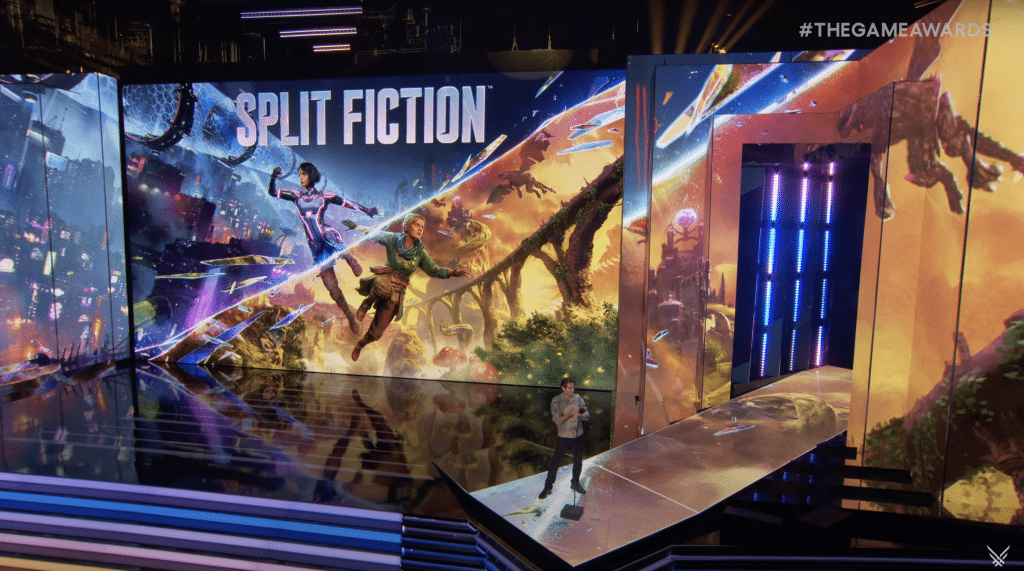 A man stands on the Game Awards 2024 stage against a background featuring 2 women dangling between sci fi and fantasy settings. The words "Split Fiction" run across the top of the artwork.