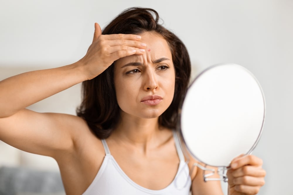 A woman is holding a mirror to her face. She is scrunching her forehead to produce fine lines. She looks displeased with the wrinkles on her forehead and holds her hand up to them to inspect the wrinkles closer.