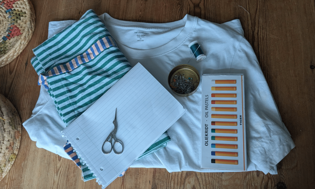 A photo of the supplies needed for the DIY. It includes a t-shirt, a green and white striped tote bag, some oil pastels, scissors, paper, needle, thread and pins.
