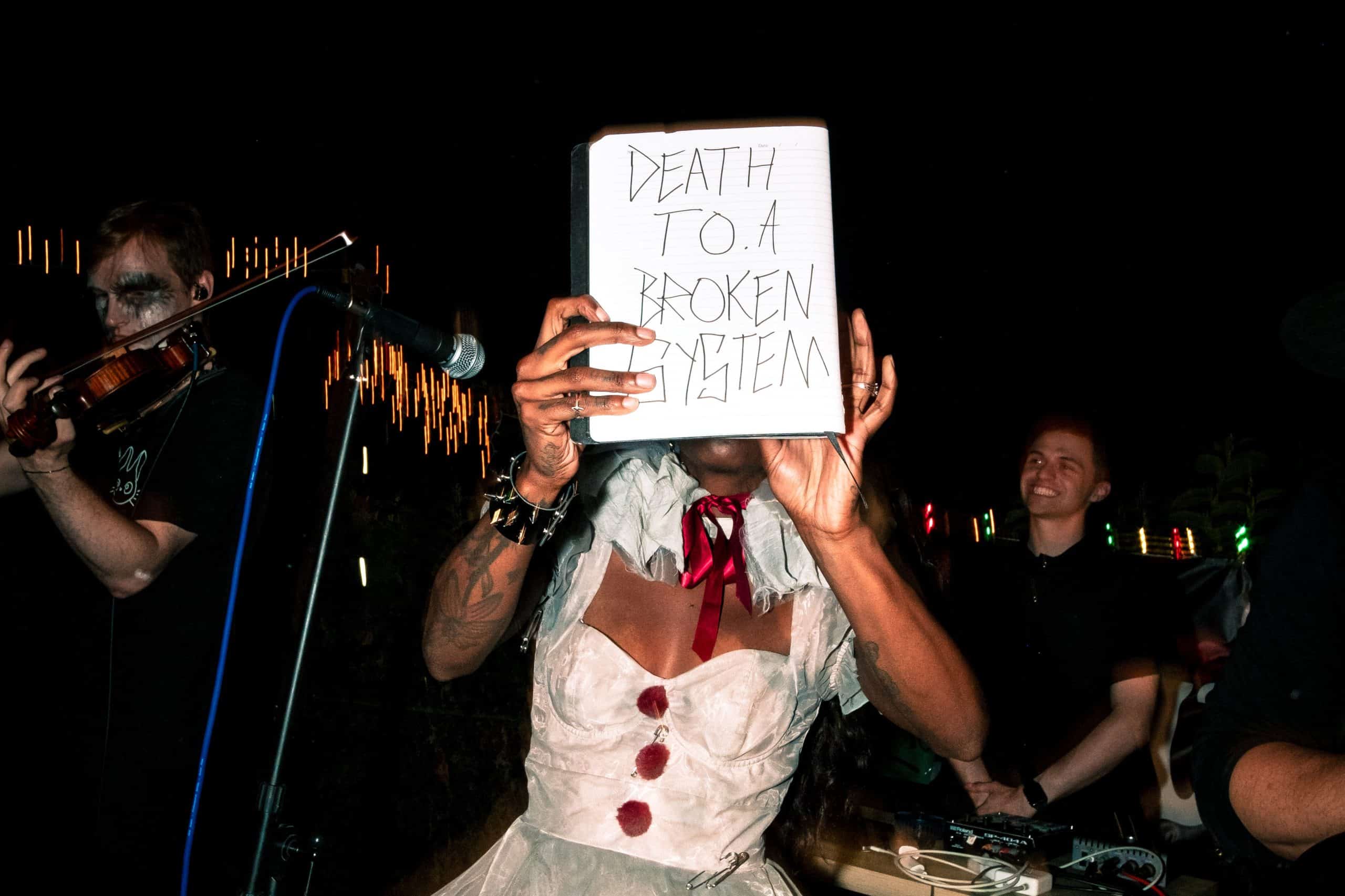 A photo of TeZaTalks holding up a sign that reads "Death to a broken system" at a music show.