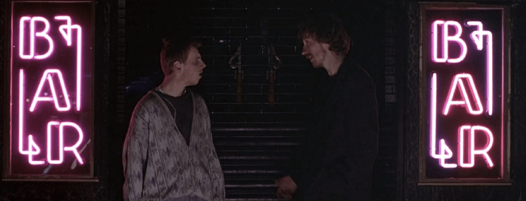 Ewen Bremer (left) and David Thewlis (right) discuss matters in the darkness in front of two pink neon "BAR" signs.