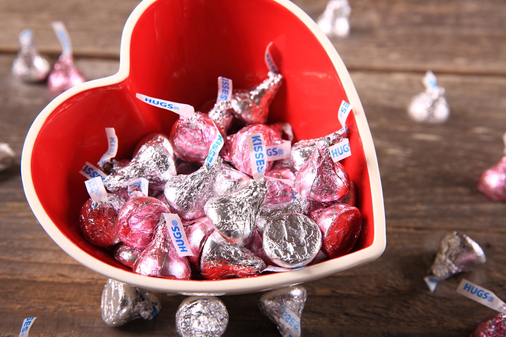 Image shows a heart-shaped bowl with silver- and pink-wrapped Hershey's kisses inside and around the outside of the bowl
