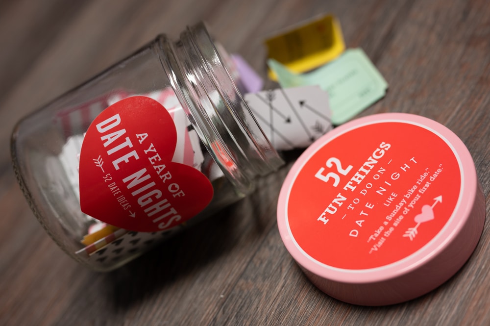 Image shows a tipped over small, glass jar. Spilling out of the jar are colorful slips of paper. The lid of the jar, face up, says, "52 Fun Things to Do on Date Night".