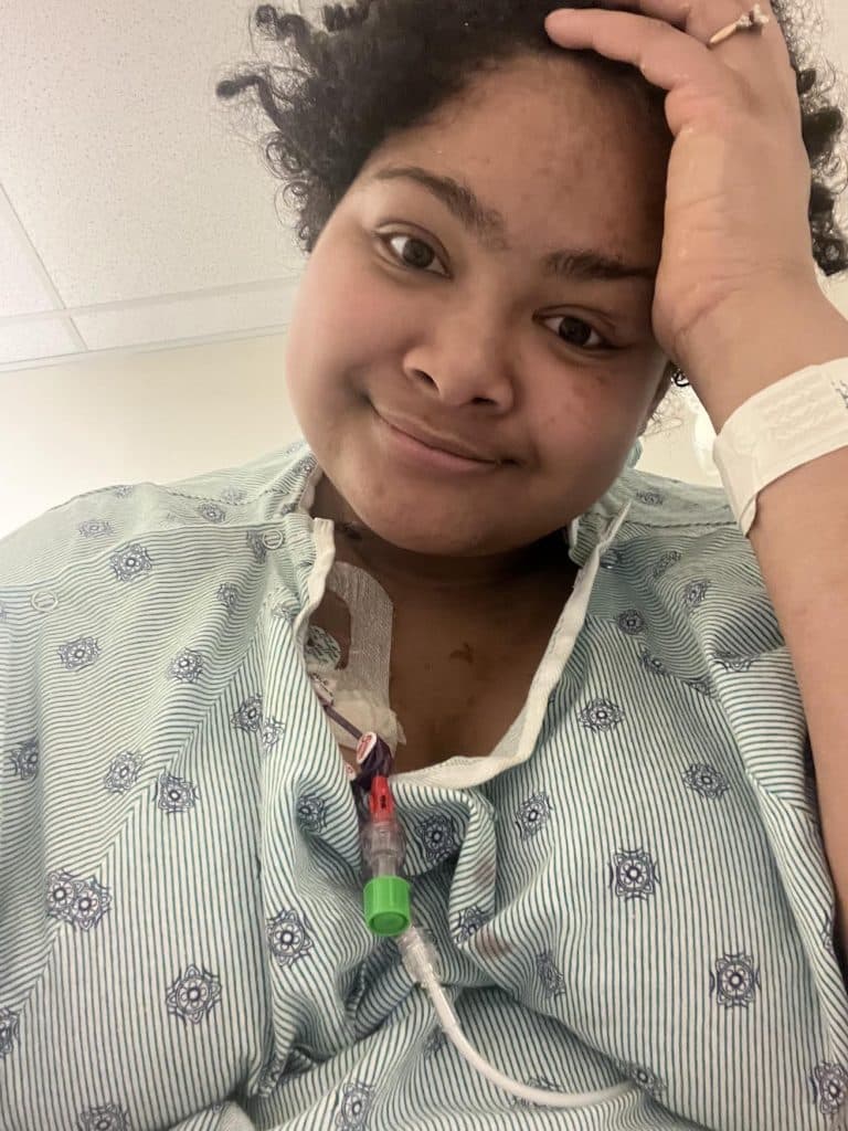 A selfie of a mixed-race woman wearing a floral-patterned hospital gown. She's resting her left hand on her head and half-smiling at the camera.