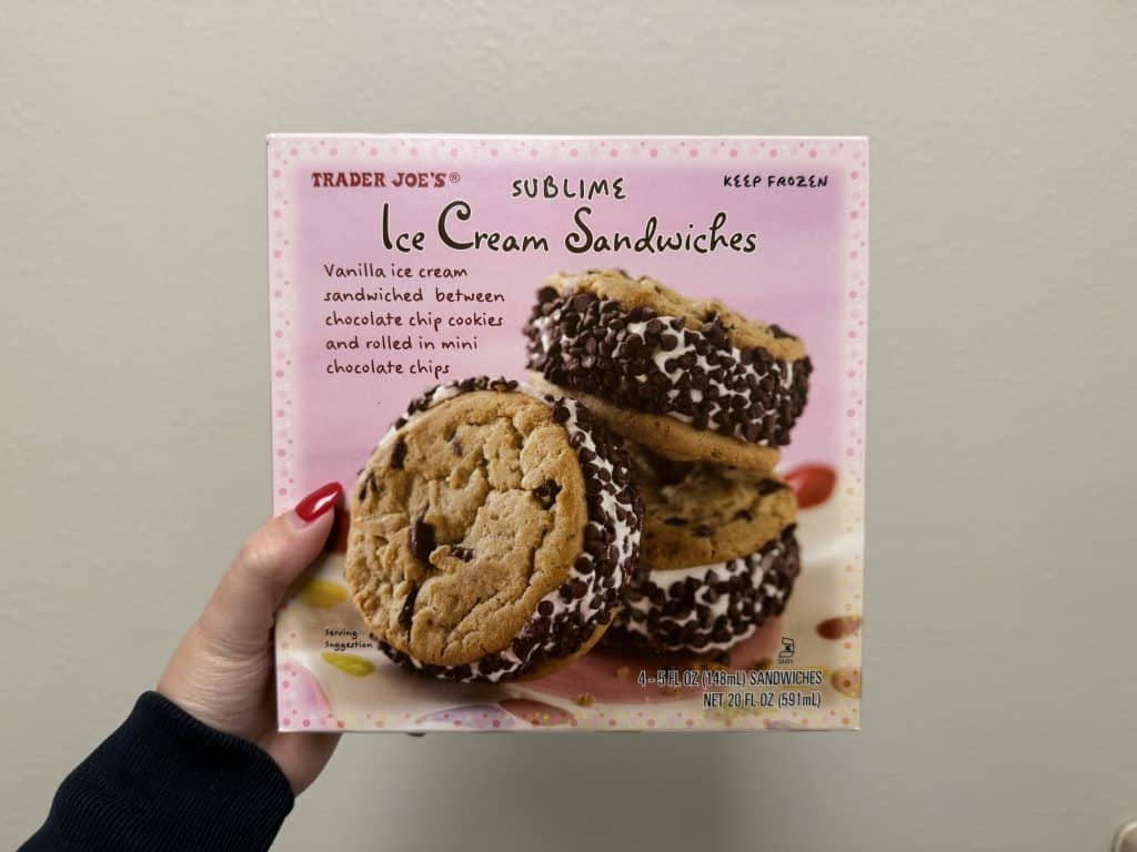 A box of frozen ice cream sandwiches. Box has baby pink background color with an image of three ice cream sandwiches displayed. Box in front of white wall background.