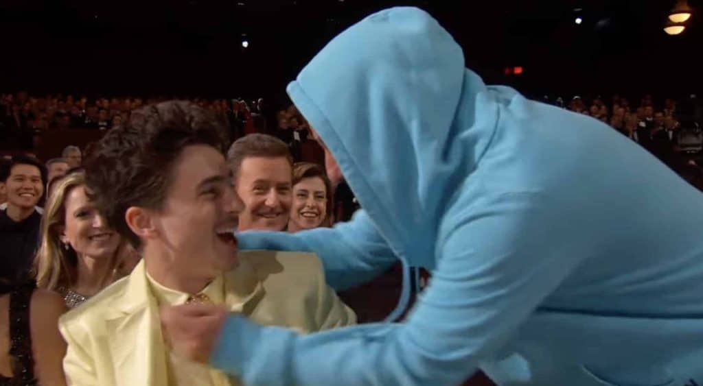 Timothée Chalamat in a yellow suit smiling at Adam Sandler in a blue hoodie.