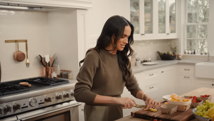 Meghan Markle is standing in a kitchen backdrop, cutting up fruit on a chopping board with a knife.