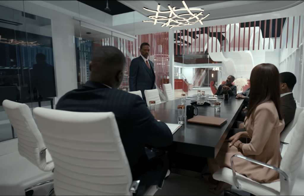The Bellaire family, sitting down at a long table for a family meeting while facing n of their family members who's standing up.