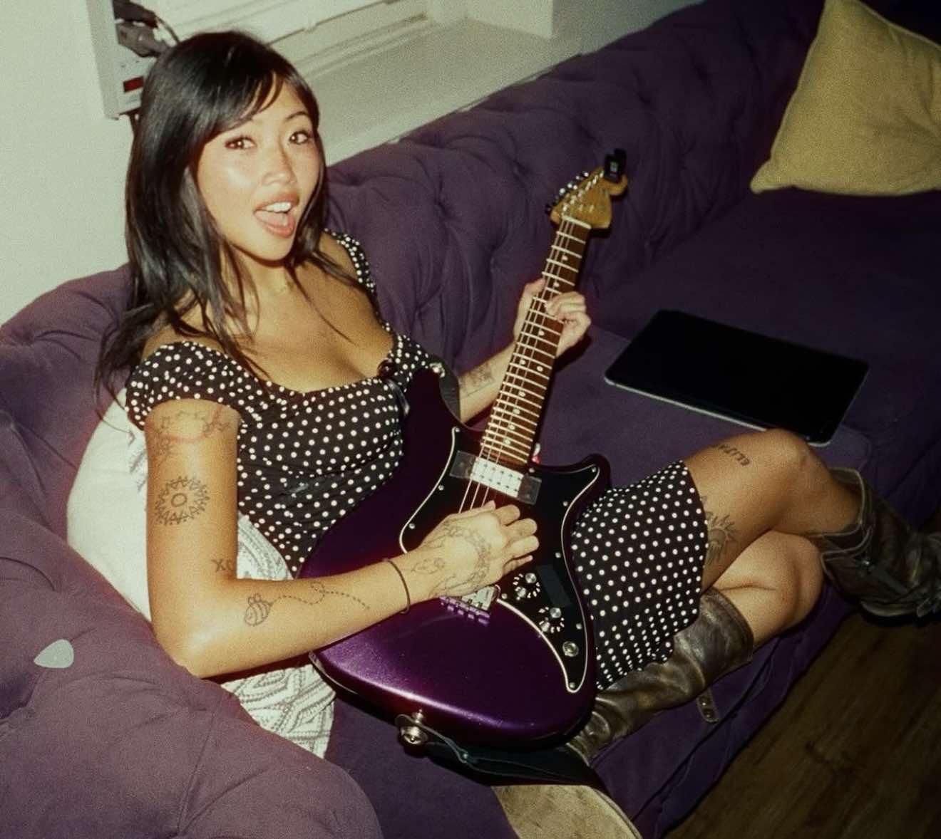 Beabadoobee laying on a couch with a purple guitar in her hands.