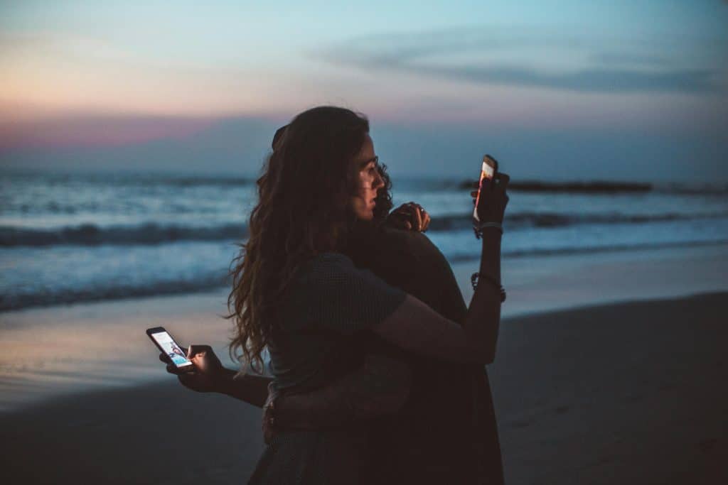 Two people hugging both looking at their phones behind the other's back