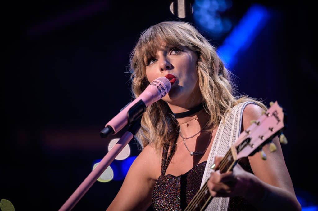 Taylor Swift looks off to the side while singing into a pink microphone and wearing red lipstick.