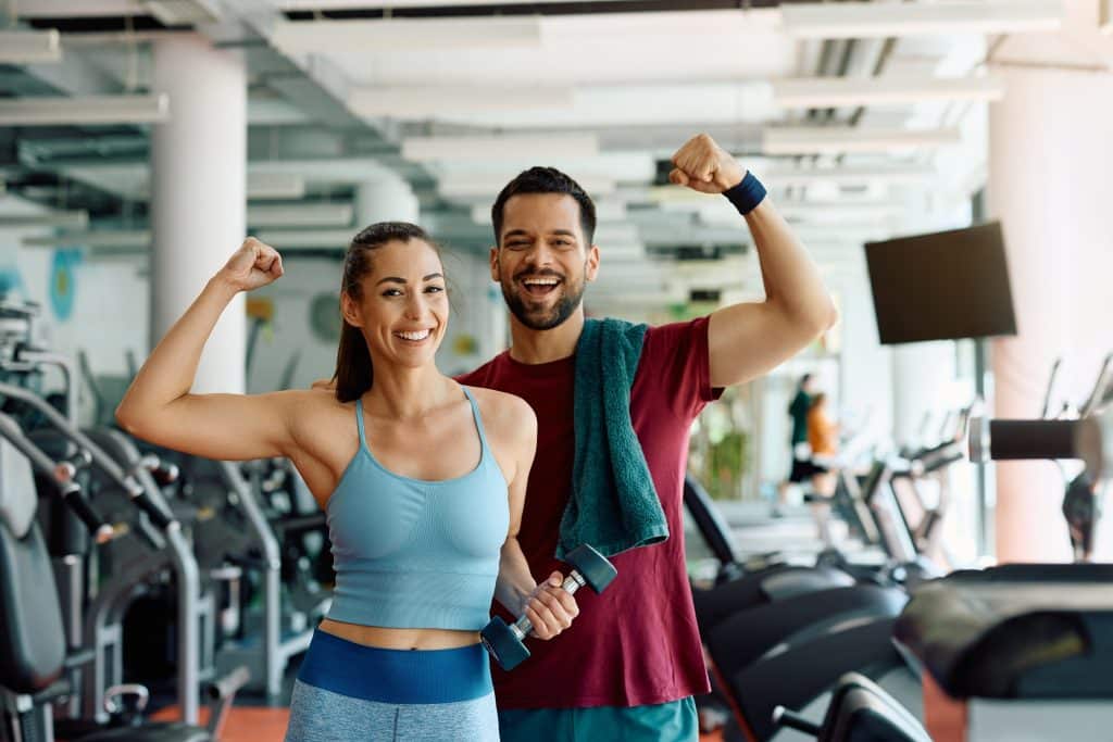 Photo shows a man and woman flexing their arms (biceps), smiling, looking at camera, in a gym setting. Gym equipment in background.