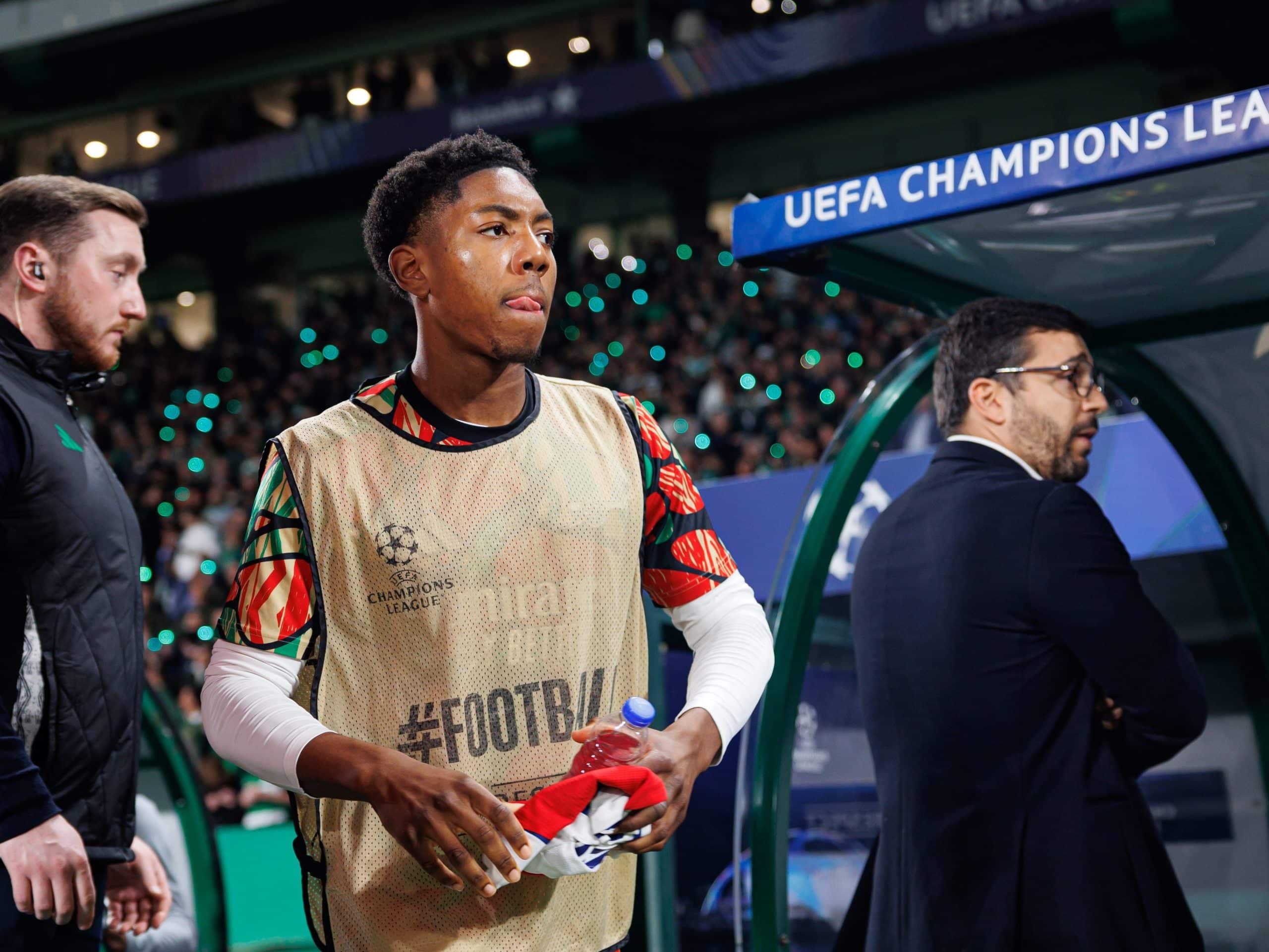Image of Myles Lewis-Skelly with a water bottle in his hands. He is on the sidelines during a football match and not playing. Two people are directly in the background.