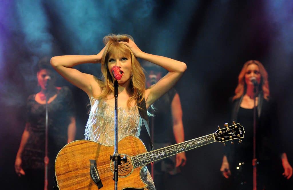 Taylor Swift sings on stage with her hands in her hair, wearing her guitar and a sparkly dress. 