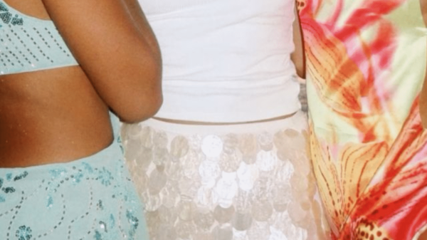 Three women dressed in vibrant Ibiza-inspired outfits pose together. The woman on the left wears a sparkling aqua blue two-piece with sequin floral designs, the center wears a white tank top with a shiny paillette mini skirt and a circular pendant necklace, and the right wears a colorful tropical satin dress in pink, orange, and green hues. The image captures a playful, summery, nightlife-ready aesthetic.