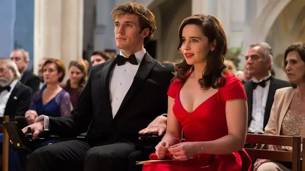 A scene from Me Before You featuring a man in a wheelchair and a woman in a red dress, sitting together in a formal setting, likely at a ceremony or event.