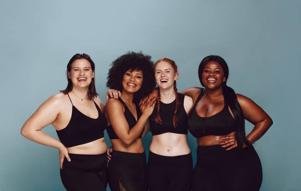 Image shows a diverse group of women, all wearing black workout sets. All women are smiling and seem to be engaged in laughter.