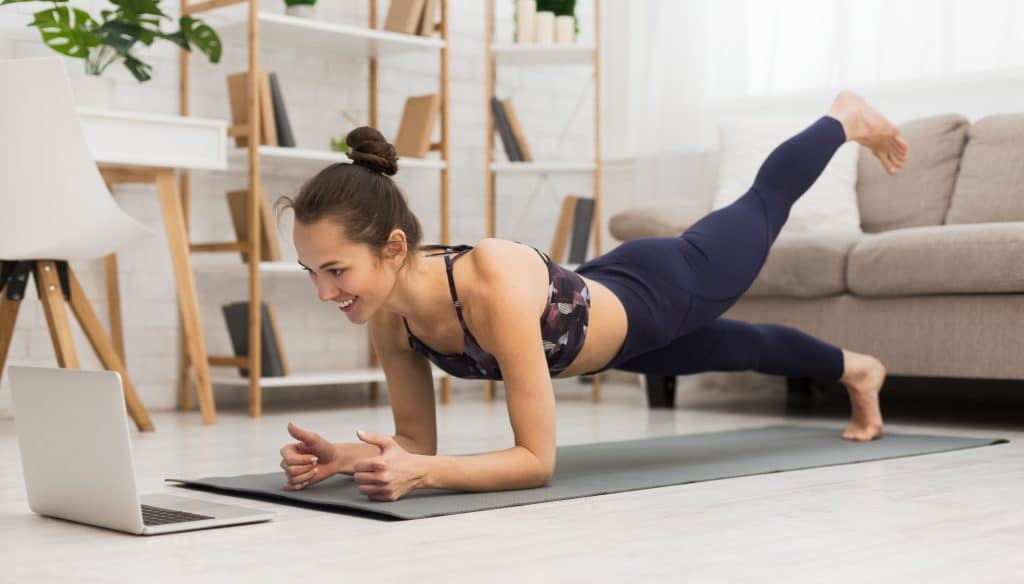Fit woman doing yoga plank and watching online tutorials on laptop, training in the living room
