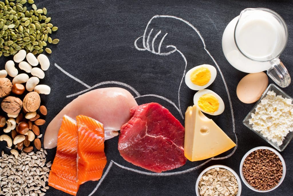 Image shows an assortment of high-protein, healthy foods sprawled out on what likes like a chalkboard type ground. A bicep is drawn on the ground with white chalk. Foods such as meats, eggs, and cheese fill up the drawn bicep.
