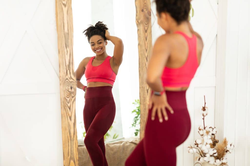 Image shows a young woman looking at herself in a full length mirror. She is wearing a workout set. She is smiling, with one hand on her head and the other on her hip.