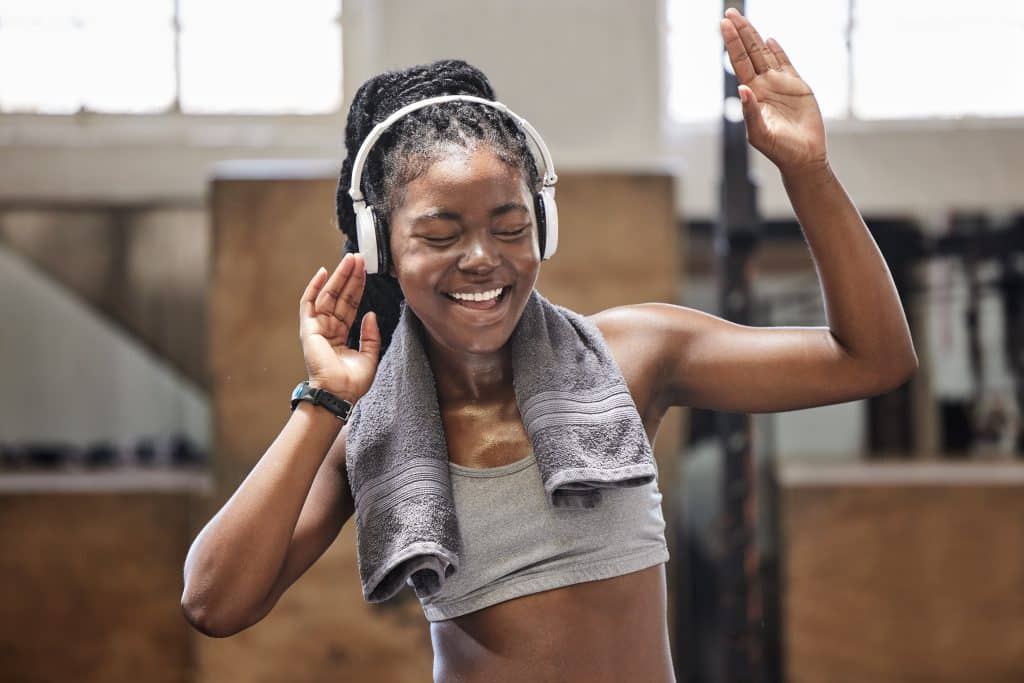 Image shows a young woman with a gray towel around her neck. She has one hand up and the other by her ear. She is wearing headphones and looks like she is singing.